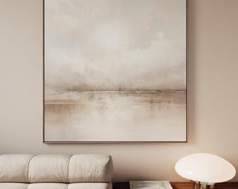 May include: Abstract landscape painting in neutral tones. The artwork features a horizontal composition with soft, blended colors of beige, brown, and white. The painting is framed in a dark brown frame and hangs on a beige wall.