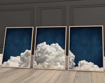 May include: Three framed prints of a white cloud against a dark blue sky. The clouds are different shapes and sizes. The frames are a light wood color.