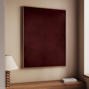 Op de afbeelding: Een ingelijst kunstwerk met een diep bordeauxrood fluwelen paneel. Het kunstwerk is ingekaderd in een dun, goudkleurig frame. Het stuk is gemonteerd op een beige muur, met een houten kast en een lamp zichtbaar op de voorgrond.