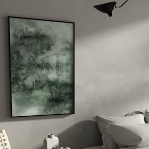 K&ouml;nnte beinhalten: Abstrakter Kunstdruck in einem schwarzen Rahmen. Das Kunstwerk zeigt eine Mischung aus Gr&uuml;n- und Graut&ouml;nen, die einen strukturierten, aquarellartigen Effekt erzeugt. Die gerahmte Kunst wird an einer hellgrauen Wand ausgestellt und verleiht eine moderne Note.