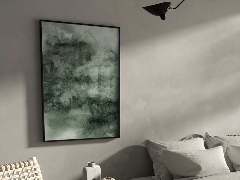 K&ouml;nnte beinhalten: Abstrakter Kunstdruck in einem schwarzen Rahmen. Das Kunstwerk zeigt eine Mischung aus Gr&uuml;n- und Graut&ouml;nen, die einen strukturierten, aquarellartigen Effekt erzeugt. Die gerahmte Kunst wird an einer hellgrauen Wand ausgestellt und verleiht eine moderne Note.
