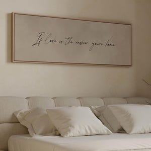 Op de afbeelding: Een beige canvas print met de tekst "If love is the answer, you're home" in zwarte cursieve letters. De print hangt boven een beige hoofdbord met witte kussens.
