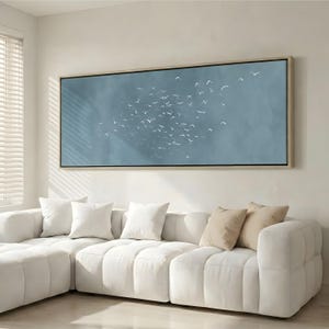 May include: A rectangular framed canvas print showing a flock of white birds flying against a light blue backdrop. The artwork hangs above a white sectional sofa with several white and beige cushions.