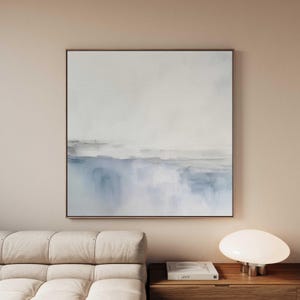 Puede incluir: Pintura abstracta de paisaje en formato cuadrado. La obra presenta una representación suave y borrosa de una línea de horizonte con tonos azules y grises sobre un fondo blanco. La pintura está enmarcada en un marco fino y oscuro.