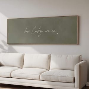 Peut inclure: Une œuvre d'art murale rectangulaire encadrée avec un fond vert sauge. L'œuvre présente la phrase manuscrite "how lucky are we" en écriture blanche, avec un petit cœur blanc. Le cadre est de couleur bois clair.