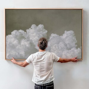 Könnte beinhalten: Ein gerahmtes Kunstwerk mit einer Wolkenlandschaft mit flauschigen weißen Wolken vor einem gedämpften grün-grauen Hintergrund. Das Kunstwerk wird von einer Person in einem weißen T-Shirt und dunklen Jeans gehalten, was die Größe des Stücks zeigt.