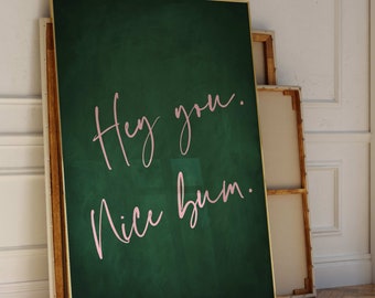 May include: A green canvas print with a gold frame featuring the text "Hey you. Nice bum." written in pink cursive.