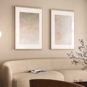 May include: Two framed abstract paintings with a beige background and subtle shades of grey and pink, hanging on a light beige wall above a cream-coloured sofa.