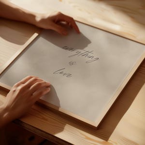 以下が含まれることがあります： 「everything is love」という言葉がエレガントな筆記体で書かれた額入りのプリント。プリントは薄いグレーの背景で、天然木のフレームに入っています。画像は木製の表面にあります。