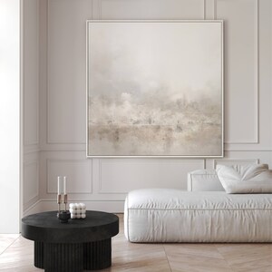 May include: A white sofa with a large abstract painting above it. The painting is in a white frame and features a light brown and grey landscape with a hint of blue. The sofa is in a modern living room with a black coffee table in front of it.