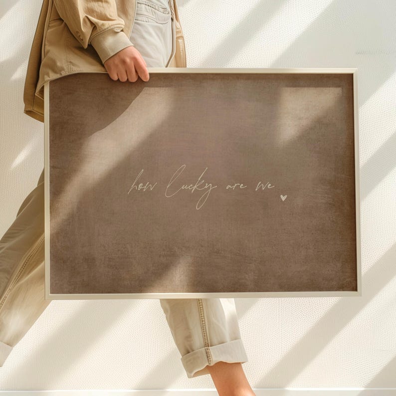 May include: A rectangular framed art print with a neutral brown background. The text "how lucky are we" is written in a delicate script, accompanied by a small heart symbol. The frame is a light, natural color.