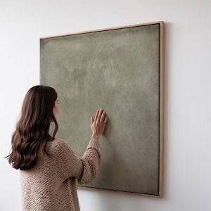 Peut inclure: Une grande œuvre d'art sur toile carrée vert olive dans un cadre en bois clair. Une personne touche la toile avec sa main. L'œuvre est accrochée à un mur blanc.