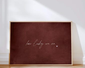 May include: A framed print with a deep red background and the handwritten text "how lucky are we" in white script. A small white heart is next to the text. The frame is a light wood color.