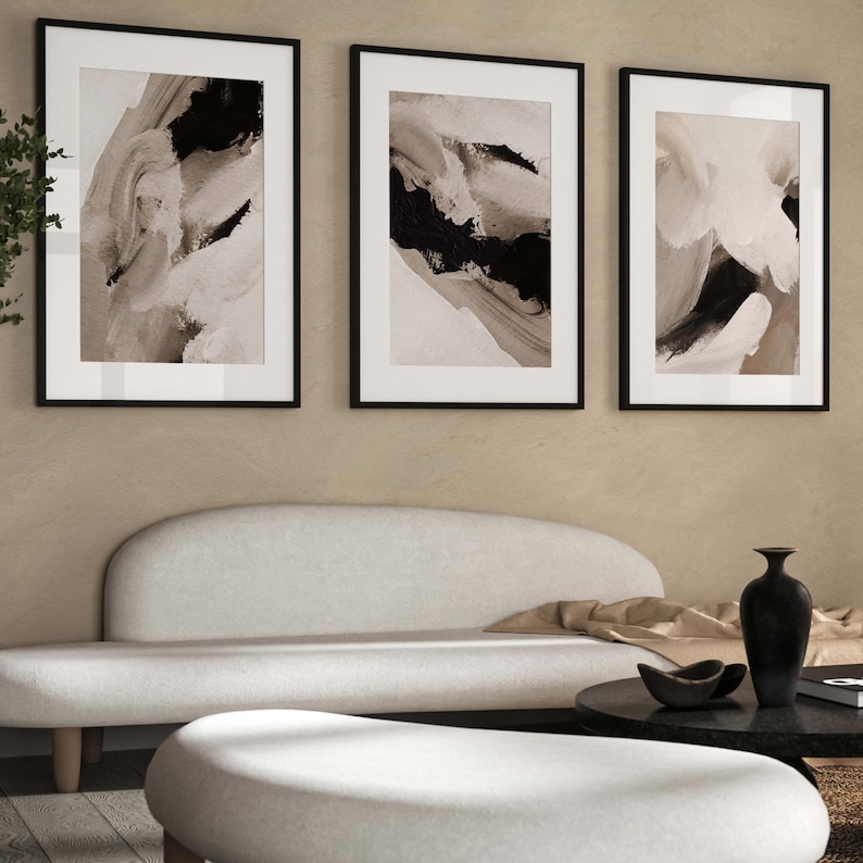 May include: Three black framed abstract art prints with white and black brushstrokes on a beige background. The prints are hanging on a wall above a white couch and a white ottoman.