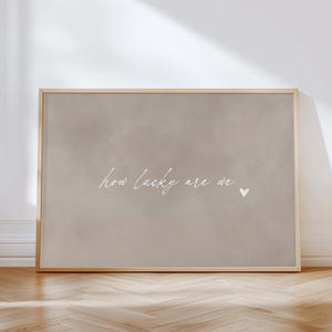 May include: A framed print with a neutral beige background. The white cursive text reads "how lucky are we" with a small heart symbol. The frame is a light wood color, and the print is displayed on a wooden floor.