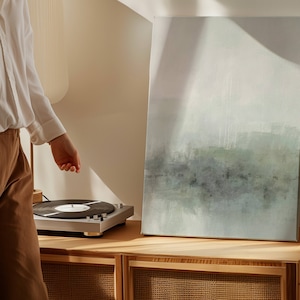May include: A silver record player with a black vinyl record on the turntable. The record player is sitting on a wooden shelf with a light blue abstract painting on the wall behind it.