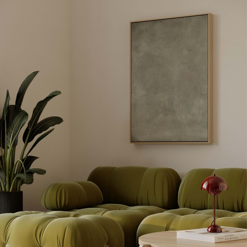 May include: A modern living room scene featuring a large olive green tufted sofa, a framed abstract artwork, and a potted plant. A small red table lamp sits on a round wooden coffee table. The walls are a neutral beige.