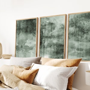 K&ouml;nnte beinhalten: Drei gerahmte Kunstdrucke mit einem ged&auml;mpften gr&uuml;n-grauen Aquarelldesign. Die Kunstwerke werden &uuml;ber einem Bett mit verschiedenen Kissen in neutralen T&ouml;nen ausgestellt. Die Rahmen sind aus hellem Holz.