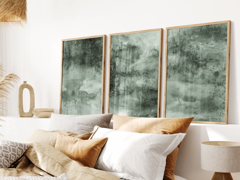 K&ouml;nnte beinhalten: Drei gerahmte Kunstdrucke mit einem ged&auml;mpften gr&uuml;n-grauen Aquarelldesign. Die Kunstwerke werden &uuml;ber einem Bett mit verschiedenen Kissen in neutralen T&ouml;nen ausgestellt. Die Rahmen sind aus hellem Holz.