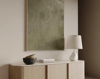 May include: A modern interior scene featuring a large olive green abstract painting in a gold frame. Below, a light wood cabinet with a marble top holds a white lamp, two bowls, and books. The room has a neutral color palette.