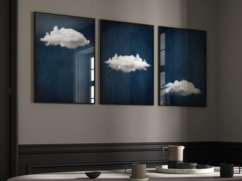 Op de afbeelding: Drie ingelijste prints met een donkerblauwe achtergrond en witte wolken. De prints zijn in een rij op een grijze muur geplaatst.