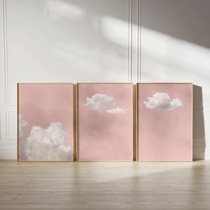 Peut inclure: Trois impressions d'art encadrées représentant des nuages. Chaque impression a un fond rose et un cadre doré. Les nuages sont blancs et duveteux, créant une esthétique douce et rêveuse. Les impressions sont exposées sur un sol en bois.