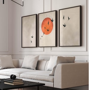 Op de afbeelding: Drie ingelijste wandkunstwerken. Het middelste stuk toont een grote oranje zon met zwarte vogel silhouetten. De andere twee stukken hebben een neutrale achtergrond met zwarte vogel silhouetten. De frames zijn donkerbruin.