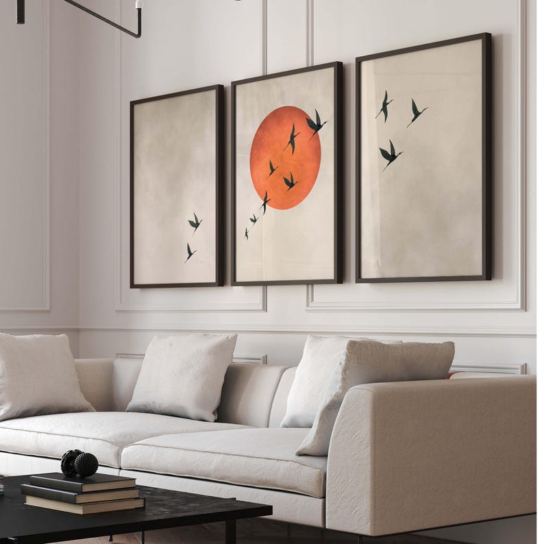 Op de afbeelding: Drie ingelijste wandkunstwerken. Het middelste stuk toont een grote oranje zon met zwarte vogel silhouetten. De andere twee stukken hebben een neutrale achtergrond met zwarte vogel silhouetten. De frames zijn donkerbruin.