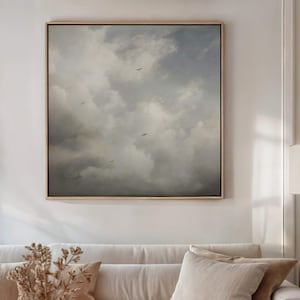 May include: A framed artwork showing a cloudy sky with birds in flight. The painting uses a palette of grey, white, and blue tones, creating a calm and atmospheric scene. The artwork is displayed above a cream-coloured sofa with decorative cushions.