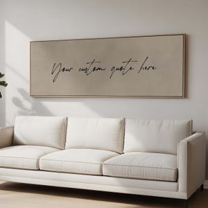 May include: A rectangular canvas art piece with a light brown background and a thin, gold-colored frame. The text "Your custom quote here" is written in black cursive script. A white sofa is below the artwork.