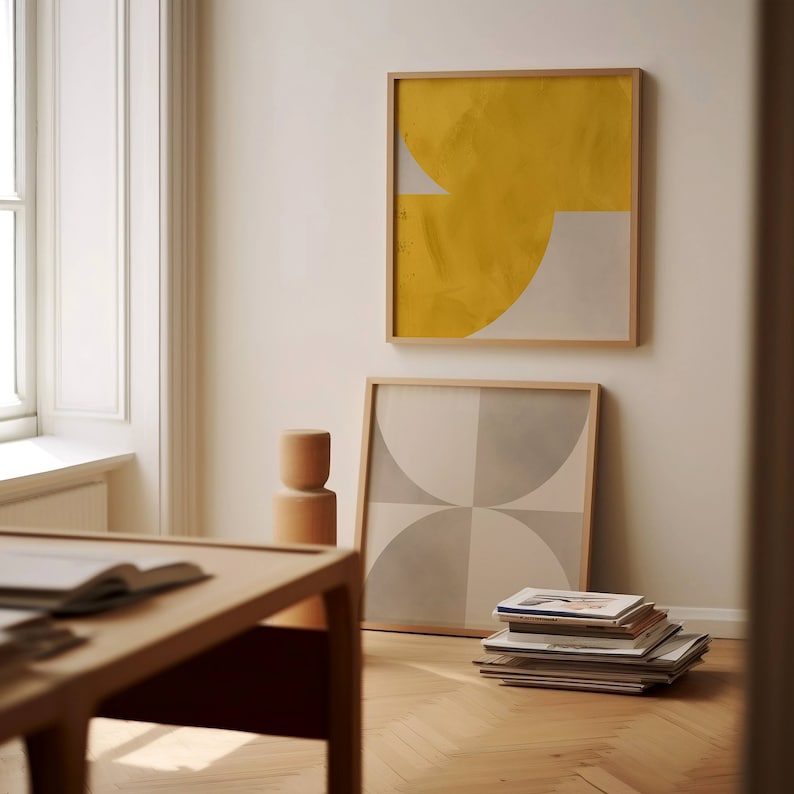 May include: Two framed abstract art prints with geometric shapes in yellow, white, and gray colors. The prints are hanging on a white wall in a room with a wooden floor and a wooden table.