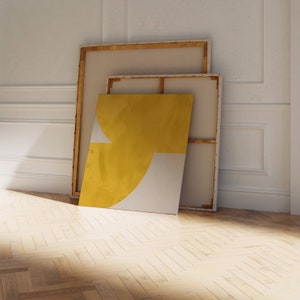 May include: Two framed canvases leaning against a white wall. The front canvas is yellow and white with a geometric design. The back canvas is beige.