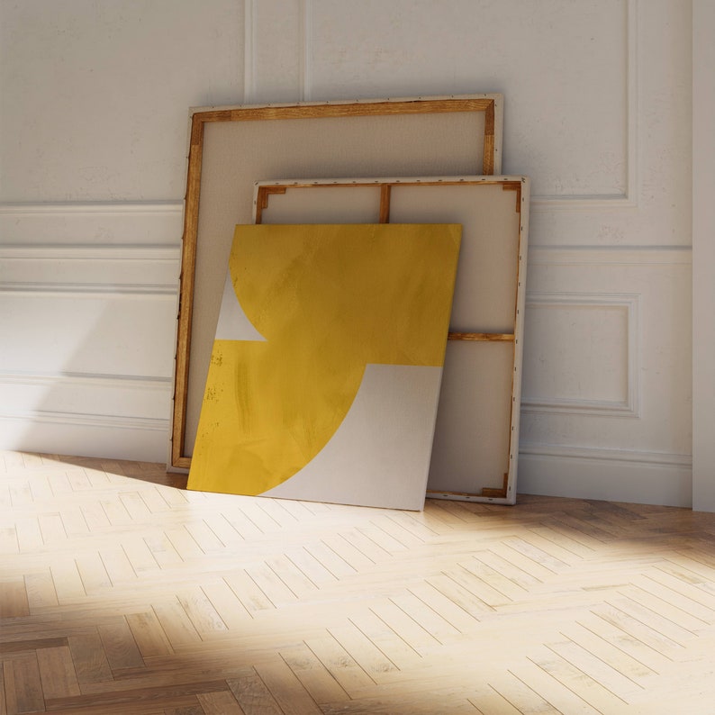 May include: Two framed canvases leaning against a white wall. The front canvas is yellow and white with a geometric design. The back canvas is beige.