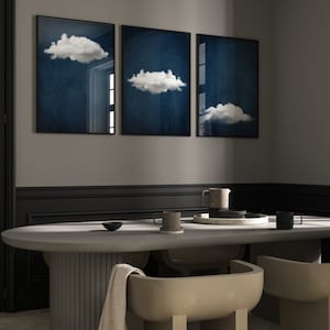 Op de afbeelding: Drie ingelijste prints van witte wolken tegen een donkerblauwe achtergrond hangen aan een grijze muur boven een betonnen eettafel met twee stoelen.