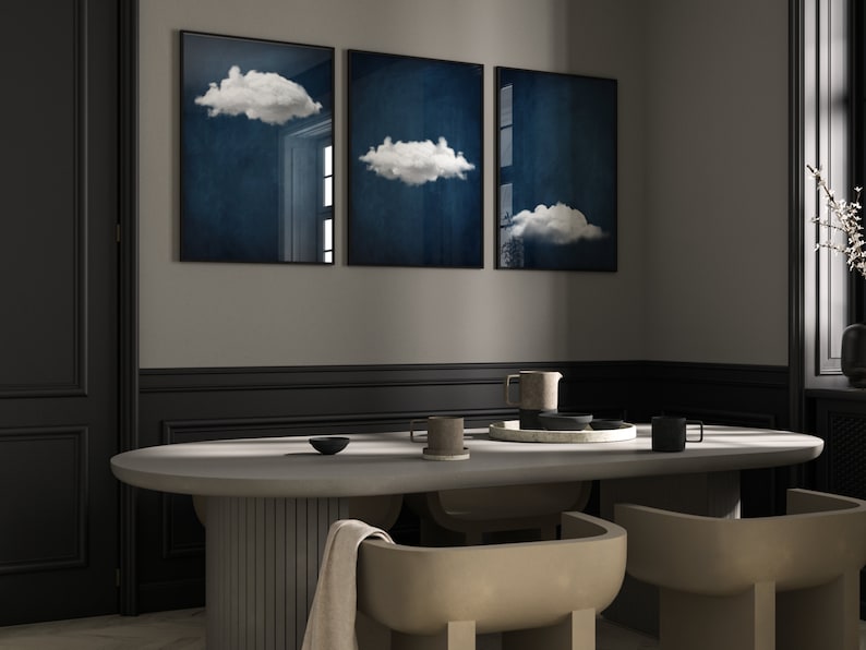 Op de afbeelding: Drie ingelijste prints van witte wolken tegen een donkerblauwe achtergrond hangen aan een grijze muur boven een betonnen eettafel met twee stoelen.
