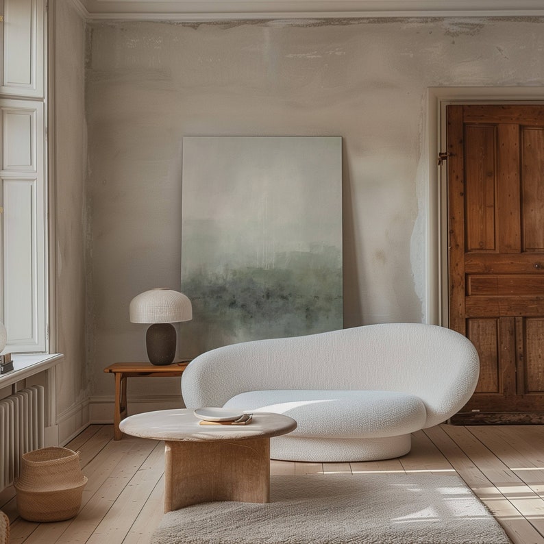 May include: A white, curved sofa with a light wood coffee table and a lamp with a cream-colored shade. The sofa is in a room with a large painting on the wall and a wooden door.