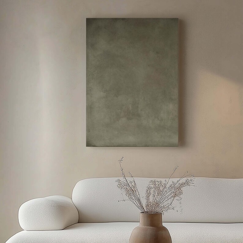 May include: A rectangular, olive-green abstract painting hangs on a beige wall. Below, a white sofa with rounded arms and a vase with dried flowers add to the minimalist decor. The painting's texture is visible.