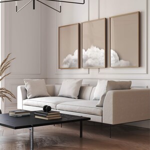 May include: A white sofa with three cushions sits in front of a wall with three framed prints of clouds. The prints are in gold frames and have a light brown background.