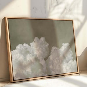 Op de afbeelding: Een ingelijste canvas print met een serene wolkenlucht. Het kunstwerk toont zachte witte wolken tegen een gedempte groen-grijze achtergrond. De print is ingelijst in een natuurlijke houten lijst, wat een warme uitstraling geeft.