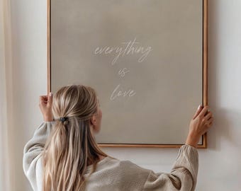 May include: A person hangs a framed canvas print with the text "everything is love" on a white wall. The canvas is a light beige color with white text.