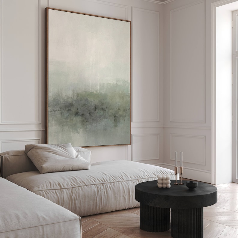 May include: A large abstract painting in shades of green and white hangs on a white wall above a white couch. The painting is framed in a thin, wooden frame. The couch is a modular sectional with a low profile and a tufted back. A black coffee table with a fluted base sits in front of the couch.