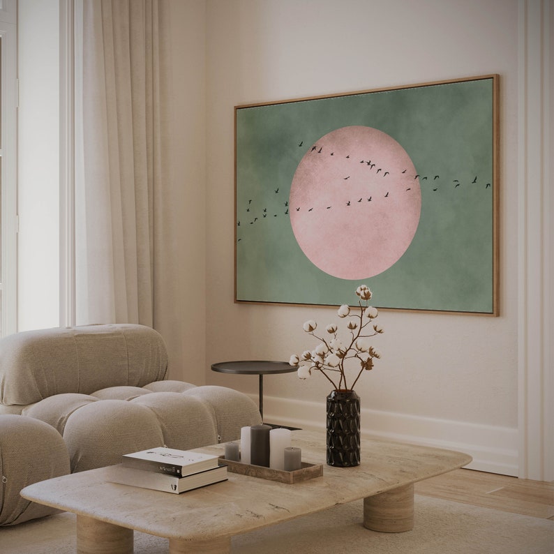 May include: A living room interior with a light beige sofa, a coffee table, a vase with white cotton flowers, and a framed abstract painting with a pink circle and black birds on a green background.