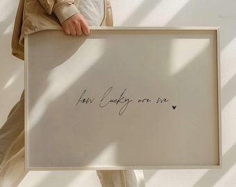 May include: A framed print with the text "how lucky are we" in a handwritten font, accompanied by a small black heart. The frame is a neutral color, and the background is a light beige.