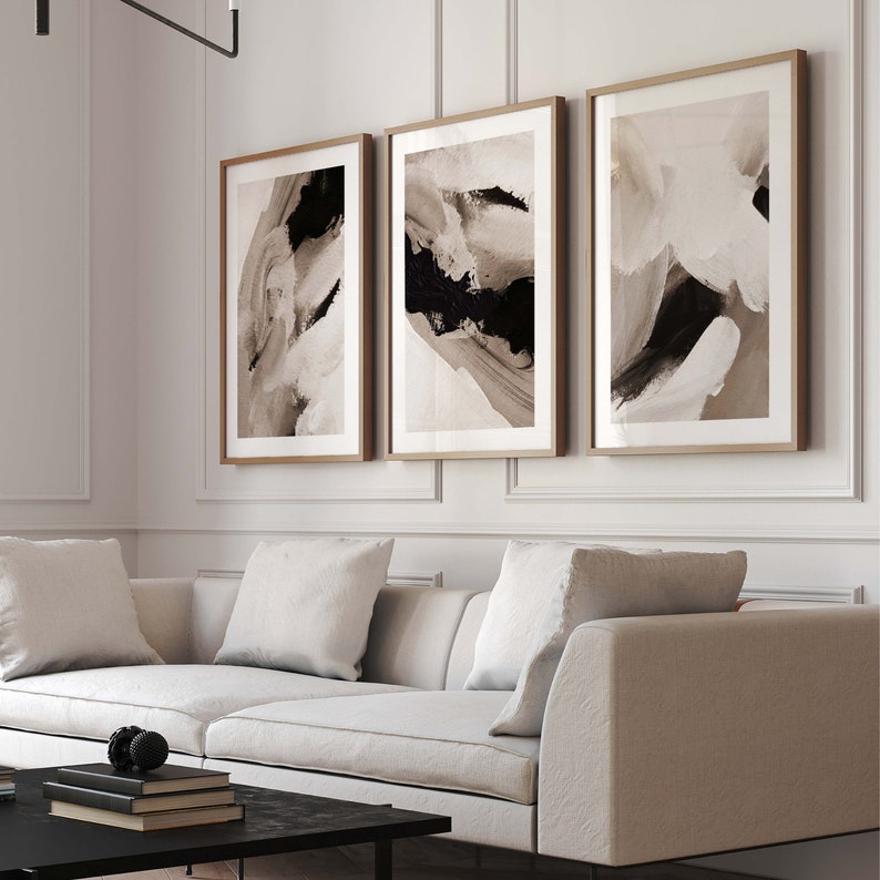 May include: Three abstract art prints in brown and black tones with a white background. The prints are framed in natural wood and are hung on a white wall above a white sofa.
