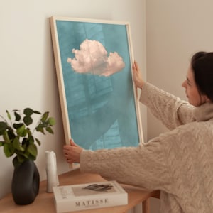 May include: A person hangs a framed print of a pink cloud on a teal background. The print is in a light wood frame.
