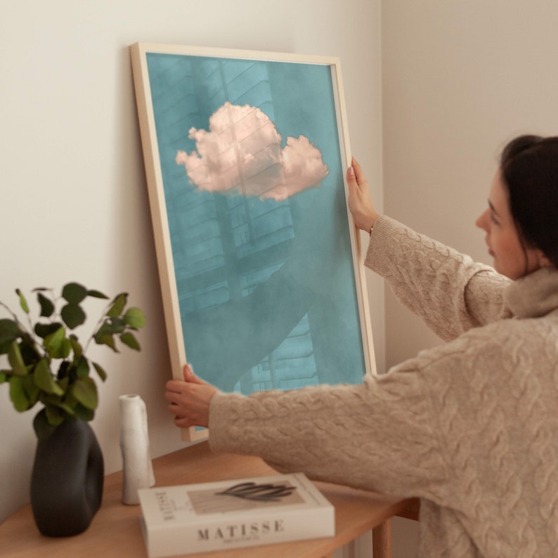 May include: A person hangs a framed print of a pink cloud on a teal background. The print is in a light wood frame.