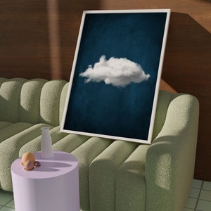 May include: A white framed print of a single white cloud against a dark blue background. The print is leaning against a light green sofa with a small purple side table and a white vase.