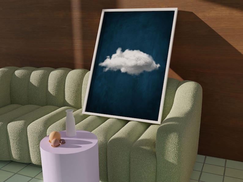 May include: A white framed print of a single white cloud against a dark blue background. The print is leaning against a light green sofa with a small purple side table and a white vase.