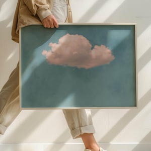 Könnte beinhalten: Ein gerahmter Druck eines einzelnen, großen, rosafarbenen Wolken vor einem blauen Himmel. Der Druck wird von einer Person gehalten.