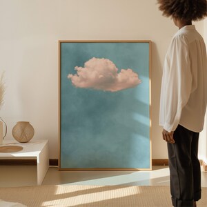 May include: A framed print of a single pink cloud against a blue sky. The print is in a gold frame and is hanging on a white wall.